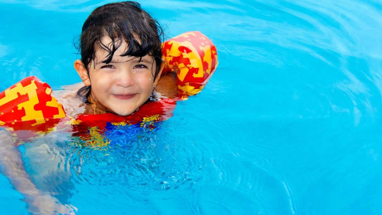 Niño pequeño con chaleco salvavidas siendo supervisado por un adulto en una piscina segura con valla perimetral.