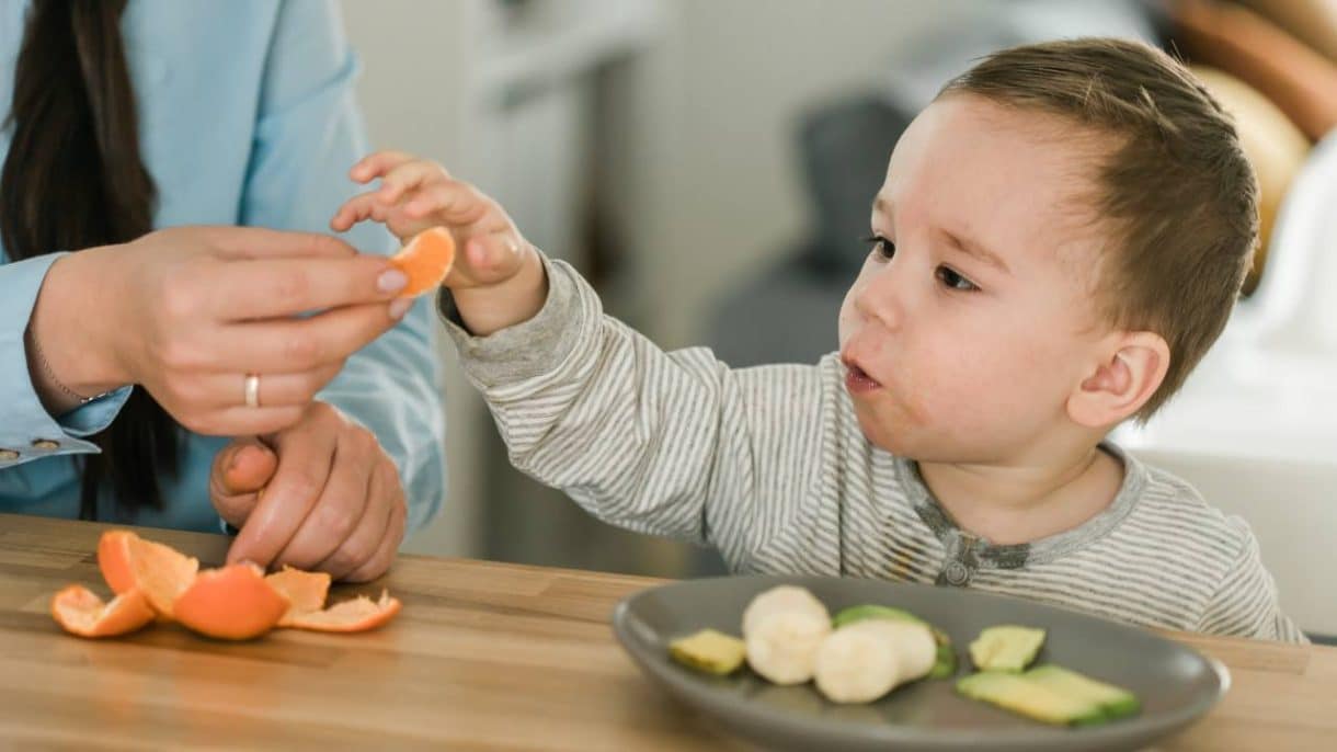 Niño pequeño comiendo fruta en trozos seguros mientras un adulto lo supervisa atentamente en la mesa.