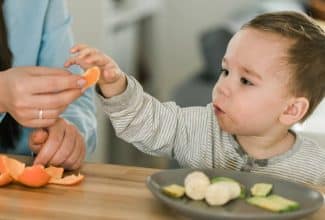 Niño pequeño comiendo fruta en trozos seguros mientras un adulto lo supervisa atentamente en la mesa.