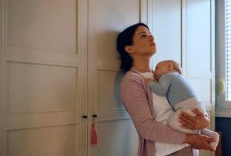 Madre con expresión de cansancio sostiene a su bebé mientras mira por la ventana, representando la depresión posparto prolongada.