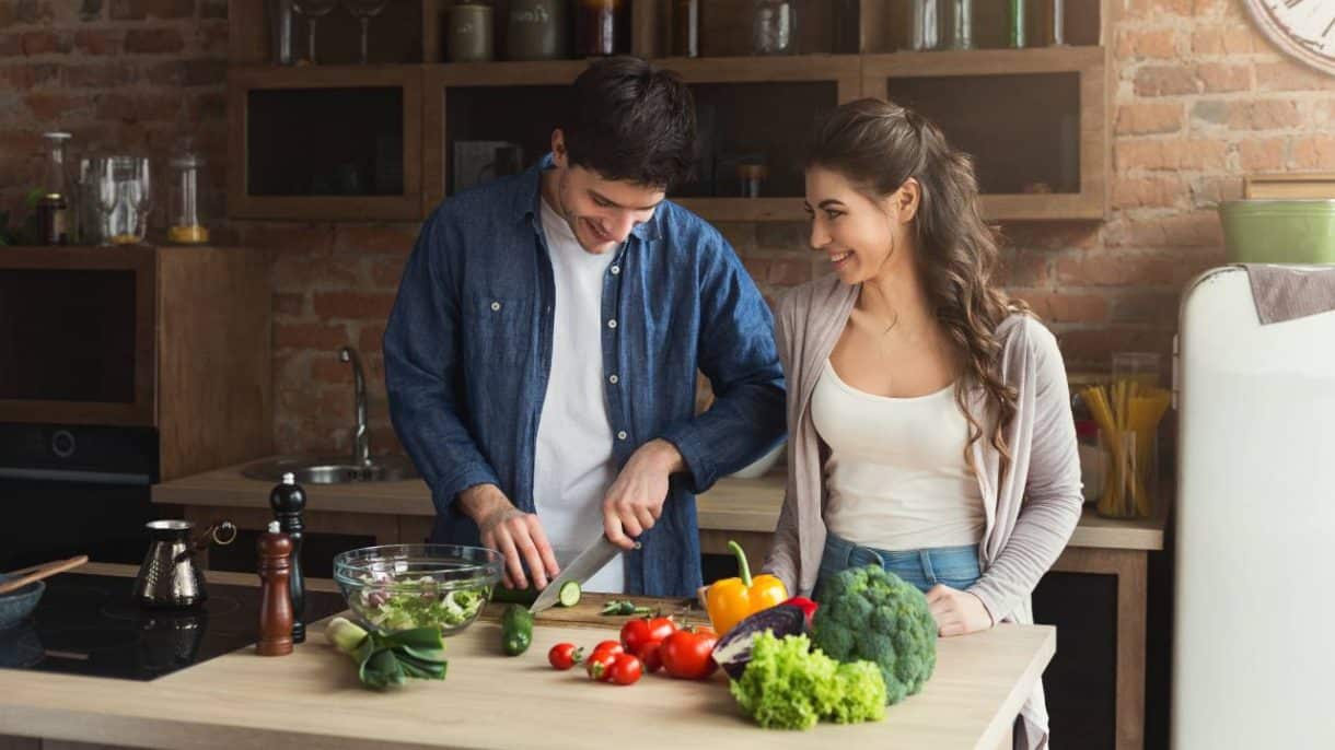 Hombre y mujer de mediana edad sonriendo mientras preparan una ensalada con vegetales frescos, representando una dieta saludable para la longevidad.