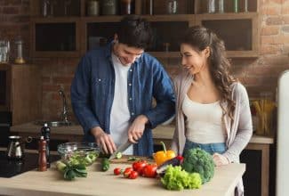 Hombre y mujer de mediana edad sonriendo mientras preparan una ensalada con vegetales frescos, representando una dieta saludable para la longevidad.